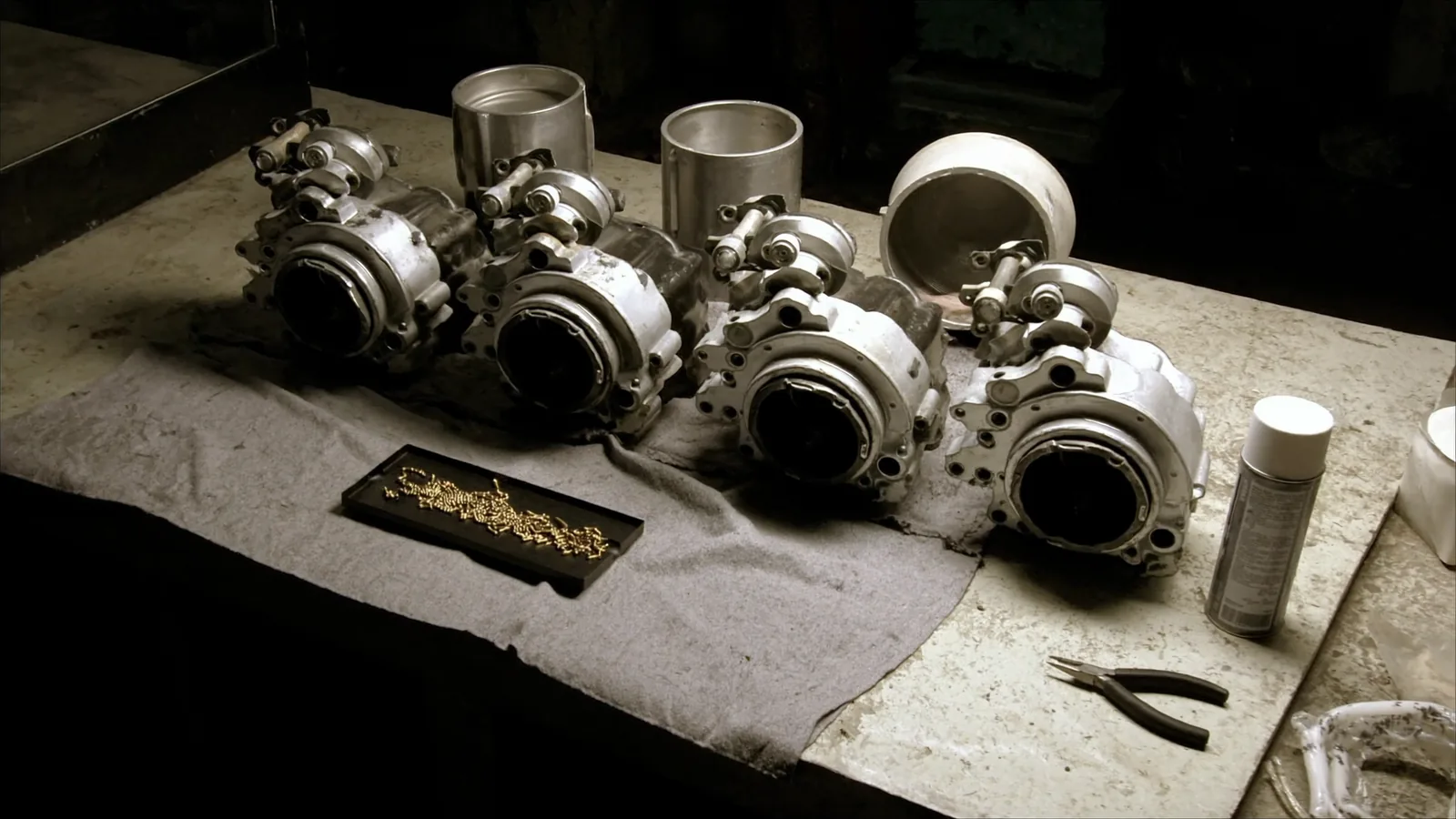 A bank of four motorcycle carburettors on a workbench cloth with the float bowls removed and lined up beside them, and small brass jets laid out on a tray.