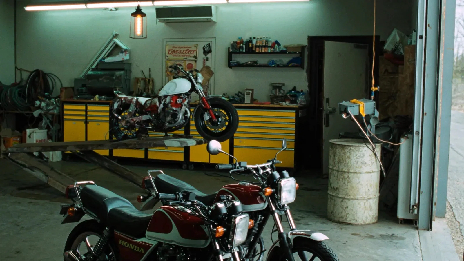 Inside a small motorcycle shop: a stripped machine up on the bench with its tank off, yellow tool cabinets behind it, and a red and white standard bike parked on the concrete by the open roll up door.