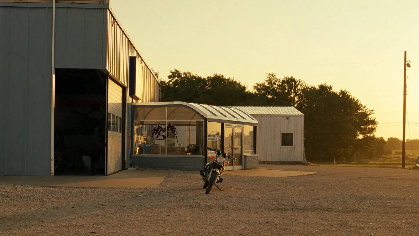 An early eighties standard motorcycle parked on gravel in low evening sun outside a grey metal shop building with the roll up door open.